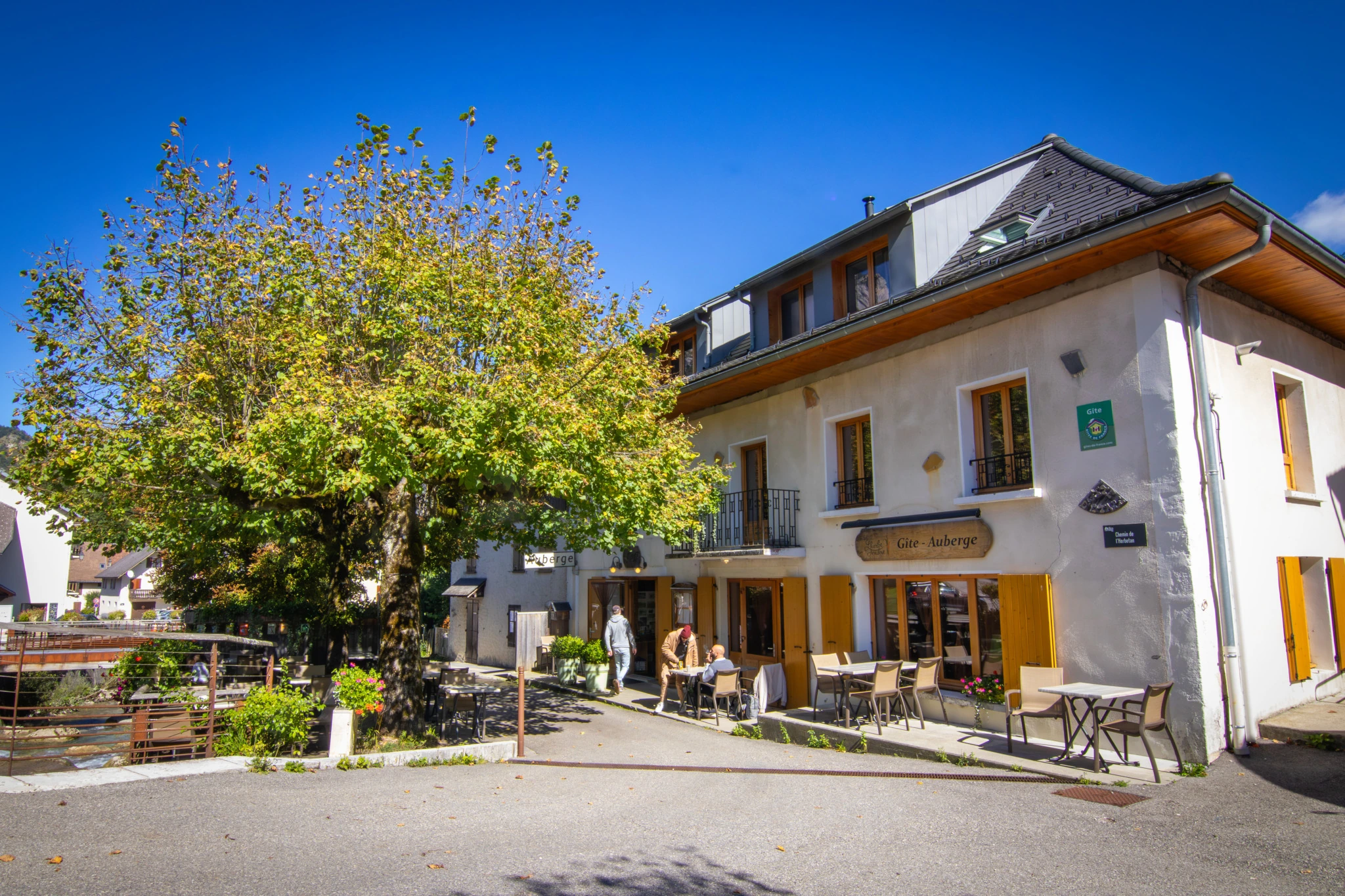 Façade extérieure du gîte-auberge L’Herbe Tendre avec terrasse, sous un ciel bleu, en Alpes Isère.